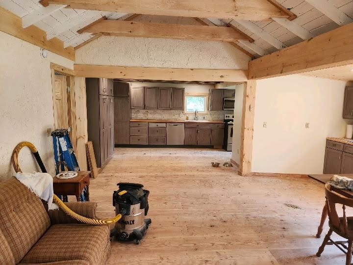 Open great room with exposed timber beams and rafters, wide pine floors, and a gray-stained kitchen with butcher-block counters