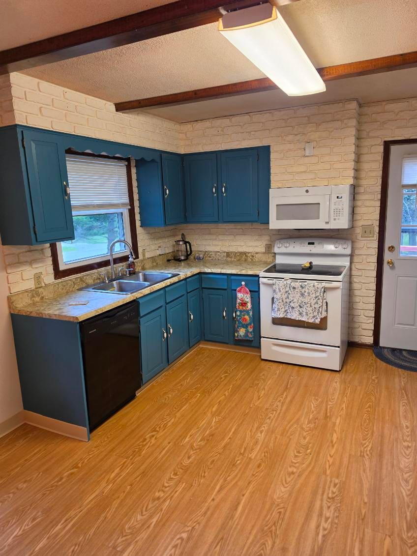 Kitchen brightened room-wide, the walls and brick backsplash painted out light behind blue cabinets, laminate counters, and a double sink under the window