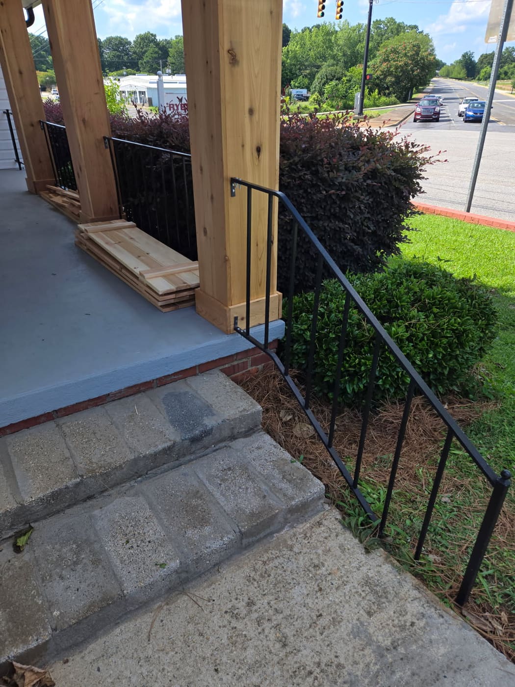 Porch post wrapped in red cedar beside an iron rail and concrete steps, with cedar stock stacked on the porch behind it