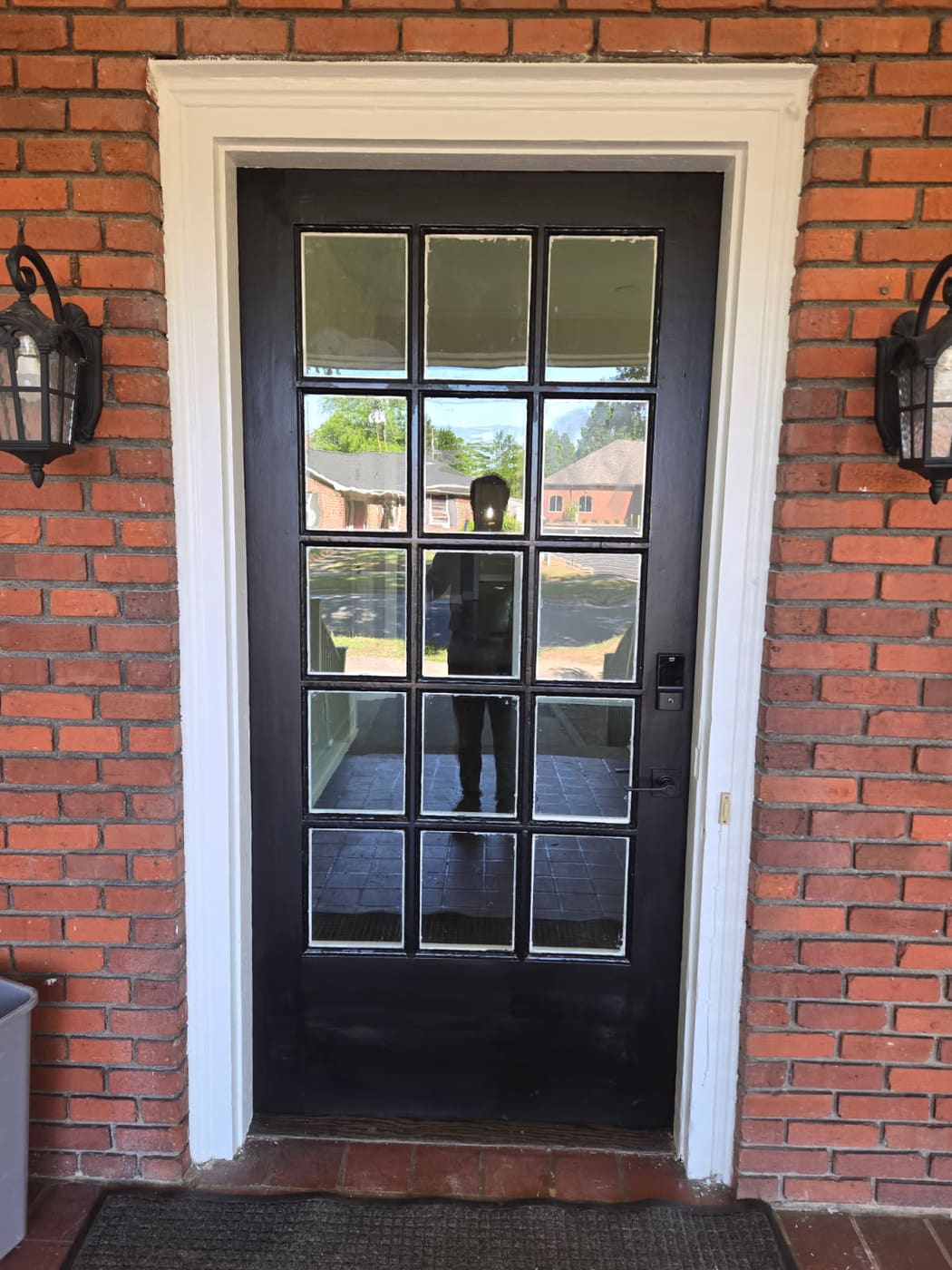 New fifteen-lite entry door hung in a brick opening, painted black with fresh white casing and a carriage lantern on each side