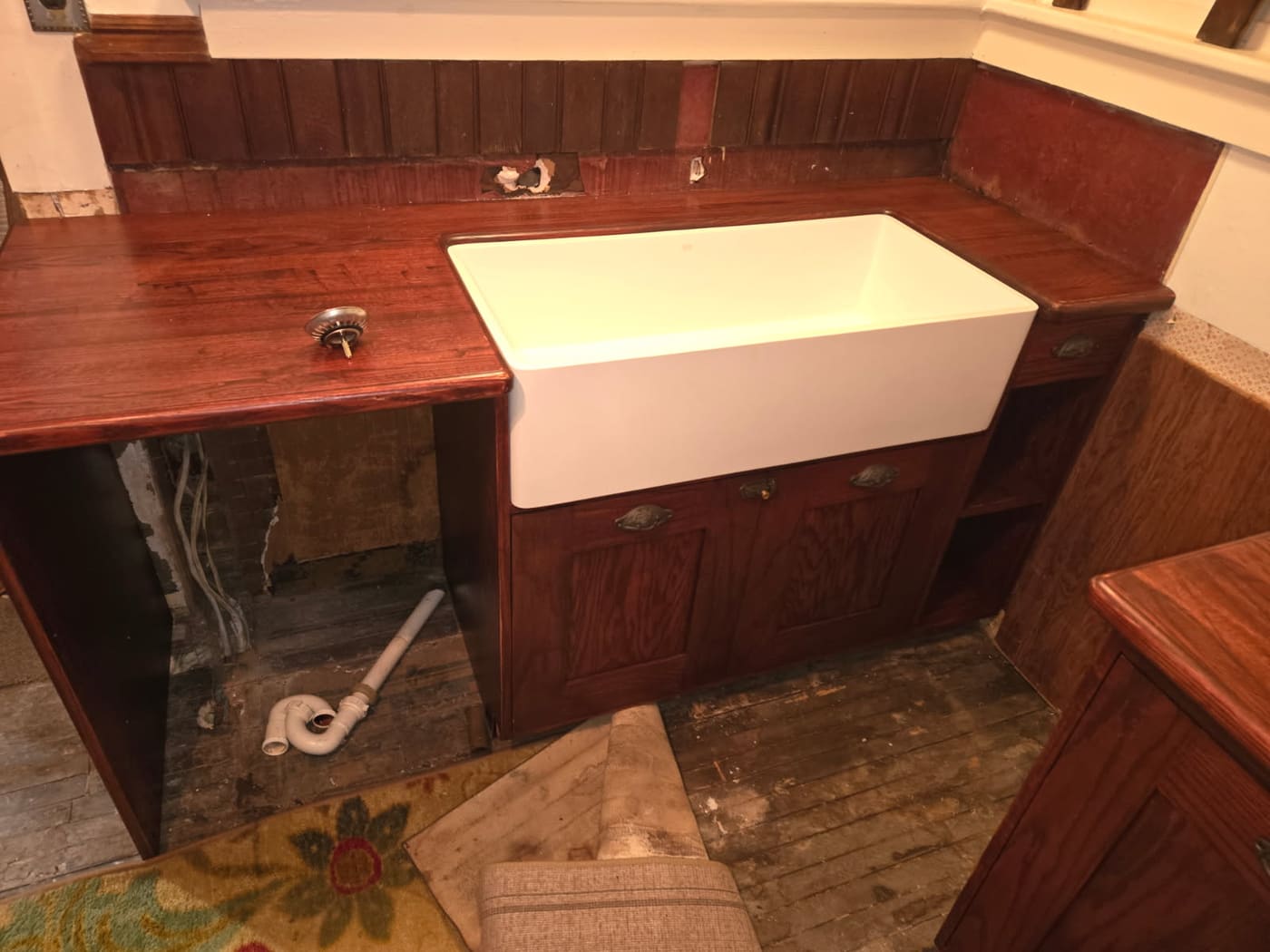 Vintage red oak kitchen cabinets built around a white apron-front farmhouse sink, photographed mid-install with the plumbing still open